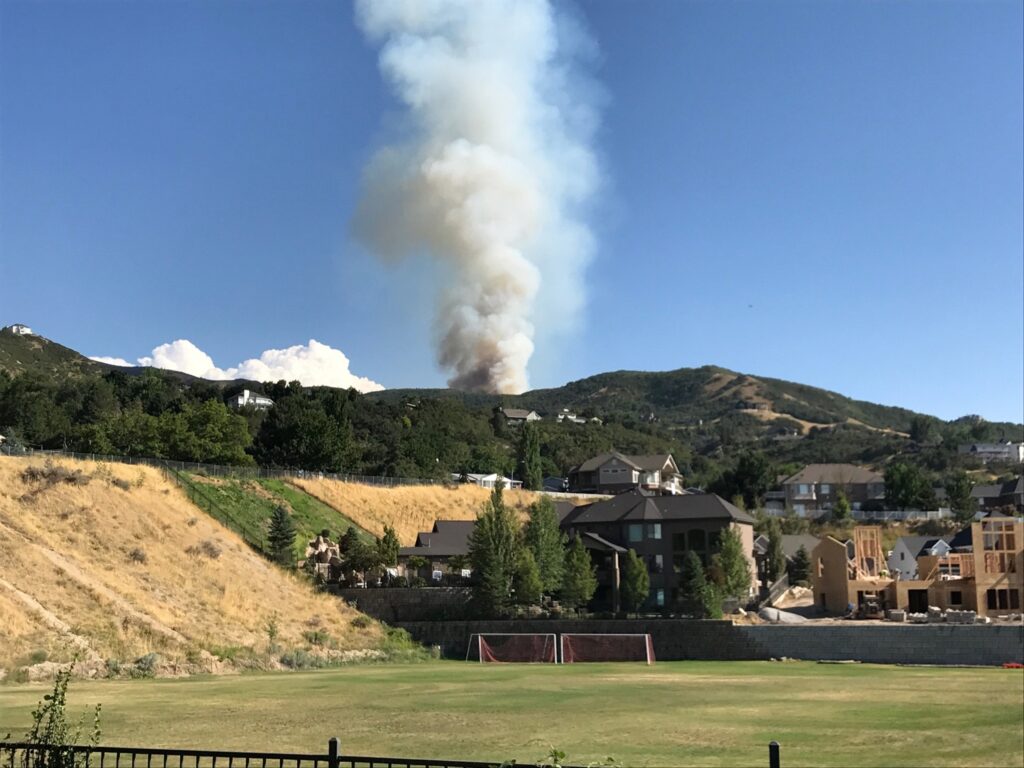 A column of wildfire smoke rises from the foothills directly above homes and a park in Bountiful.